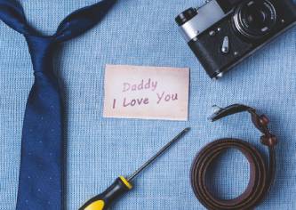 Ein Notizblock mit der Aufschrift "Daddy I Love You" liegt neben einer Kamera, einem Schraubenzieher und einer Krawatte auf einem blauen Hintergrund.